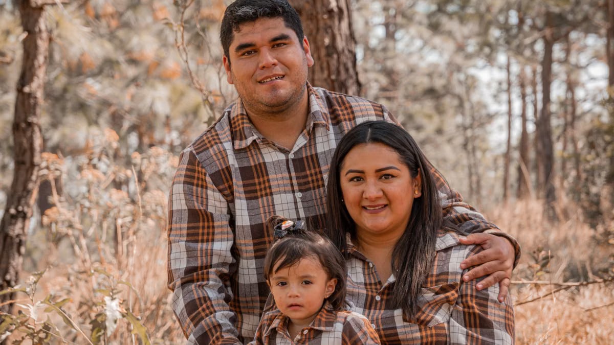 A family of three posing together in a forest wearing matching plaid shirts.